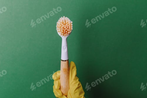 Preview: Gloved hand holds cleaning brush with wood handle against green background.