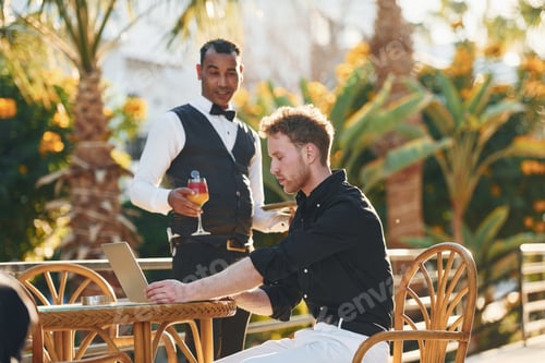 Preview: Serviced by waiter. Young man is outdoors at sunny daytime. Concept of vacation