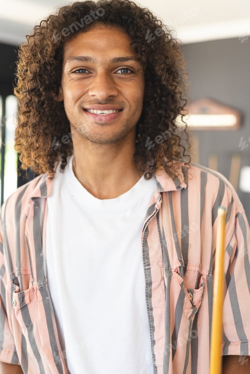 Preview: Young biracial man with curly hair smiles warmly, holding a pool cue