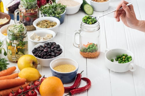 Preview: cropped view of woman adding green peas in glass jar on wooden white table