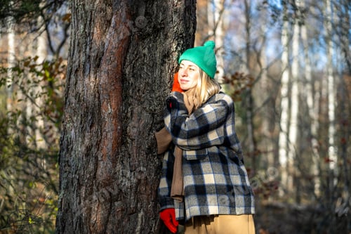Preview: Happy female traveler leaned against tree trunk with dreamy expression stands in sunny autumn woods