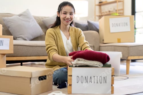 Preview: Smiling woman folding clothes for donation, organizing items with donation boxes