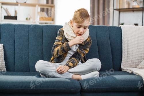 Preview: Pleasant tired sick teen boy with scarf around neck, sitting on soft couch and coughing