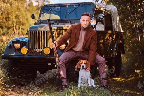 Preview: Hunter in elegant clothes together with his beagle dog next to a retro military car in the forest.