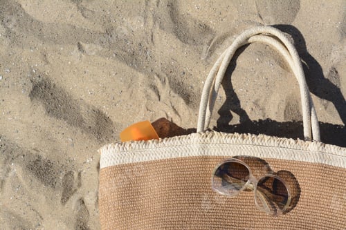Preview: Straw bag with sun protection product and sunglasses on sand, top view. Beach accessories