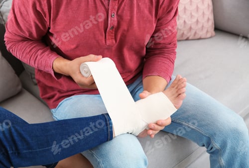 Preview: Young man applying bandage on woman's injured leg at home, closeup. First aid