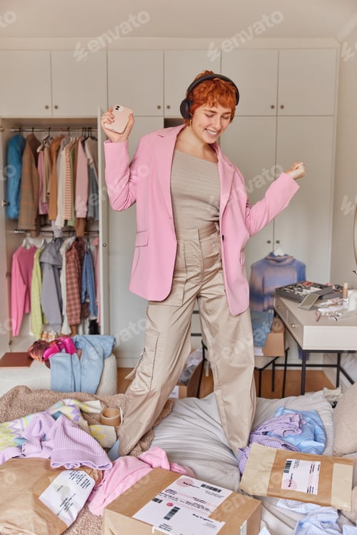 Preview: Vertical shot of happy ginger woman enjoys favorite playlist holds smartphones wears stereo