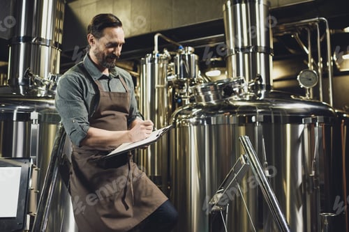 Preview: Male worker in apron inspecting industrial equipment and making notes at the brewery