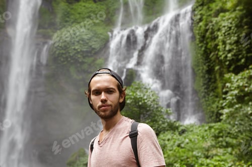 Preview: Close up portrait of handsome man wearing baseball cap backwards relaxing outdoors, standing by wate