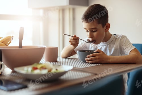 Visualização: Caucasian small teen boy eating cereals with milk and drinking tea at table in morning.