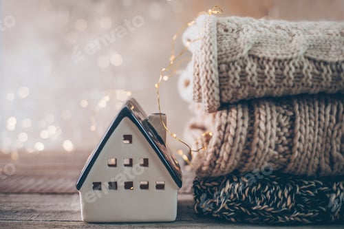 Preview: Stack of cozy winter knitted sweaters, ceramic house on blurred background with bokeh. Warm Concept