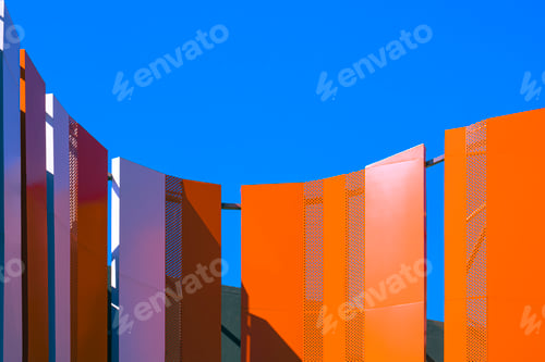 Preview: Orange and white curved steel wall outside of high building against blue clear sky background
