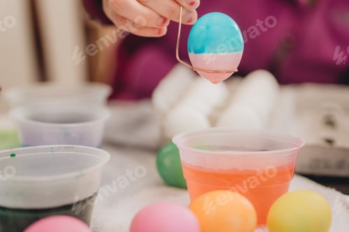 Preview: Young millennial Mother at home helping daughter paint colorful Easter eggs for playing and eating