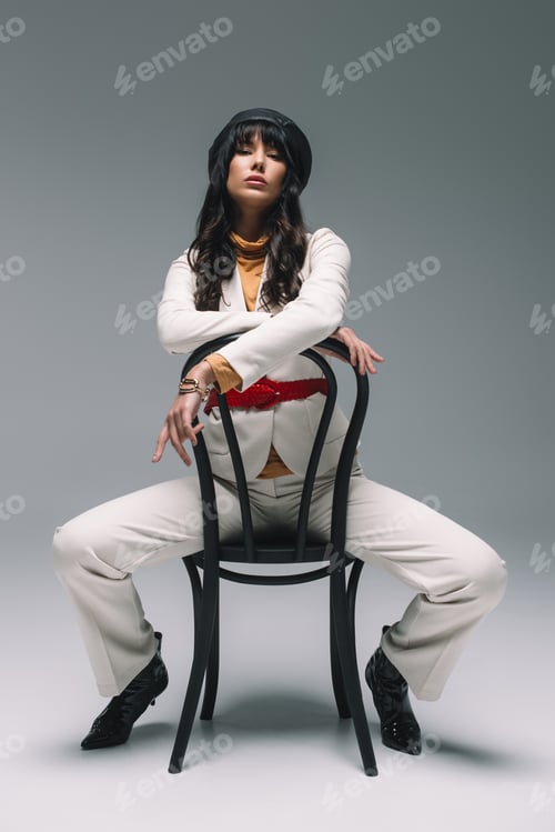 Preview: beautiful brunette woman in white suit sitting on chair and looking at camera on gray