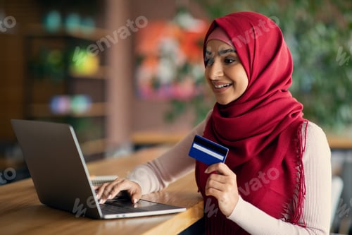 Preview: Arabic lady holding credit card, using laptop at cafe