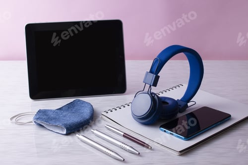 Preview: Top view of a digital tablet with headphones, notebook, and medical mask on a pink background