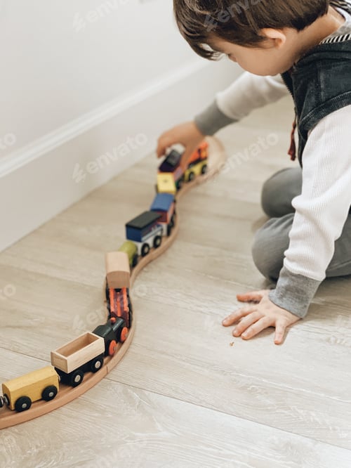 Preview: Young boy playing with a colored wooden train set on a light weathered wood floor