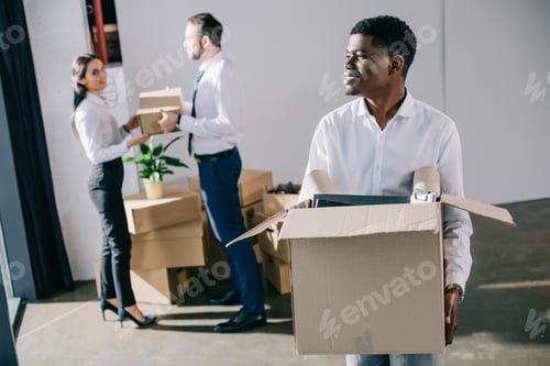 Preview: smiling african american businessman holding cardboard box while relocating with colleagues in new
