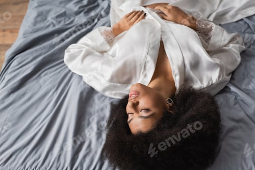 Preview: high angle view of brunette african american woman with closed eyes lying in white robe on grey