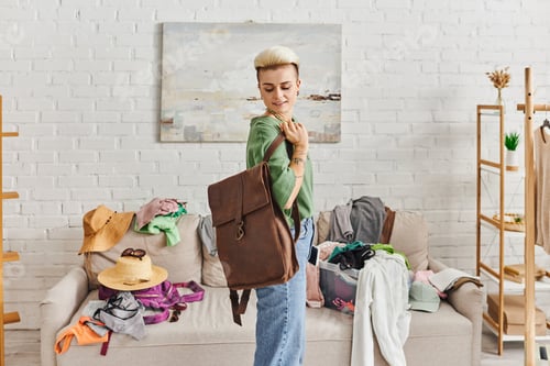 Preview: smiling woman with tattoo and trendy hairstyle standing with leather bag near couch with straw hats