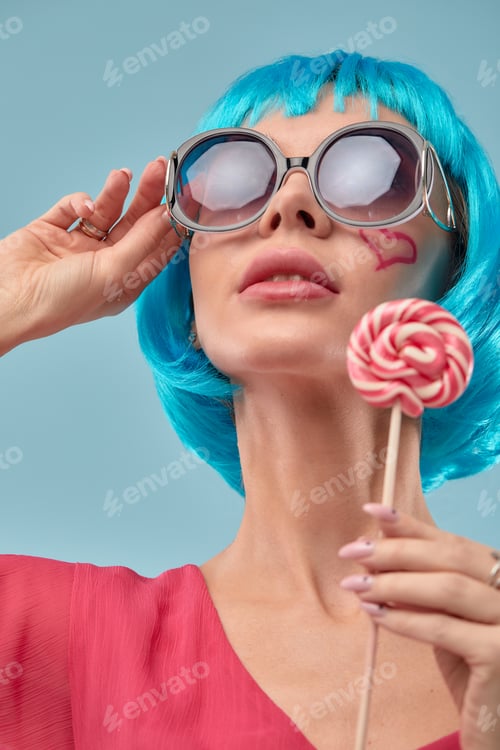 Preview: Young funky girl in blue wig and eyeglasses, holding lollipop candy, looking aside with dreamful