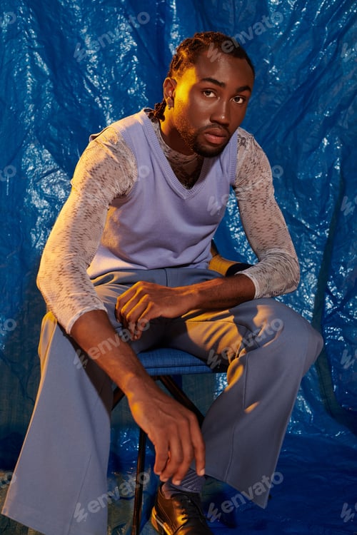 Preview: Handsome young man seated confidently against a vibrant blue backdrop showcasing urban style