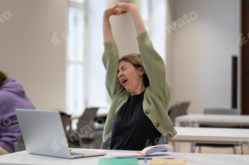 Preview: Tired female mature student taking break stretching arms while studying online in university library
