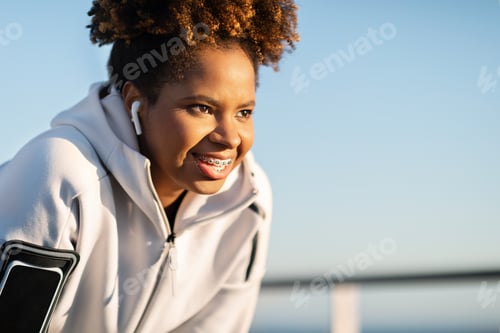 Preview: Smiling Black Woman Wearing Earphones And Armband Taking Break While Jogging Outdoors
