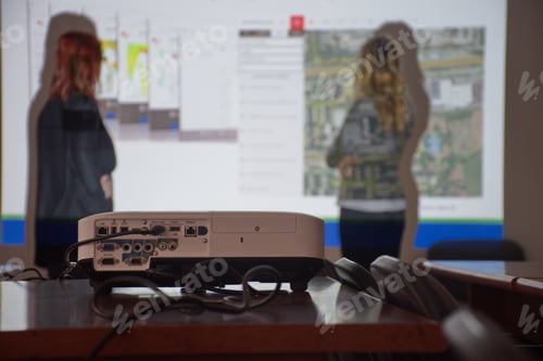 Preview: Closeup shot of a projector and two women giving a presentation