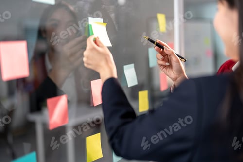 Preview: Businesswoman planning project using adhesive notes on glass wall in office