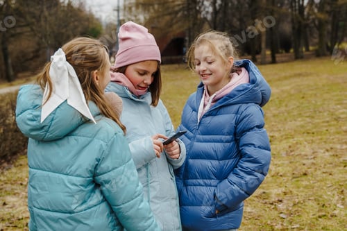 Preview: Teen girls in jackets and jeans watch smartphone, listen to music player in winter city park.