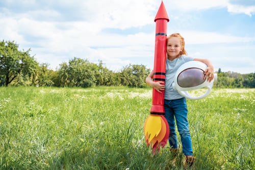 Preview: smiling red hair kid with rocket and astronaut helmet standing in summer field