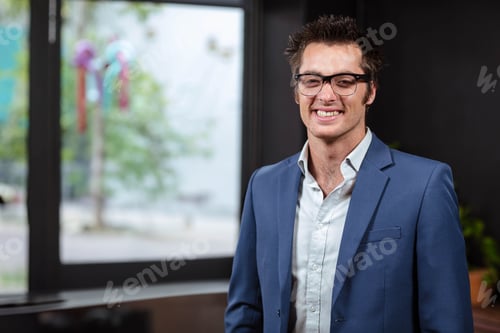 Preview: Man standing in office by glass window with ribbons wearing glasses and suit jacket, copy space