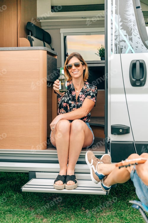 Preview: Woman showing beer bottle having fun with friends sitting at the door of their camper van