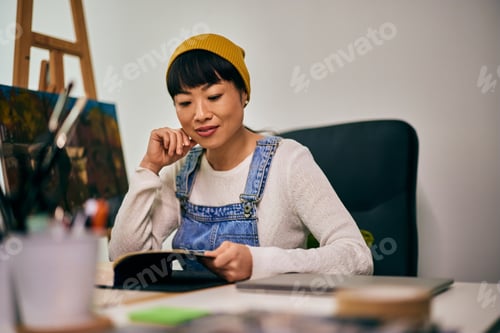 Preview: Asian woman reading a book, sitting in her art studio.