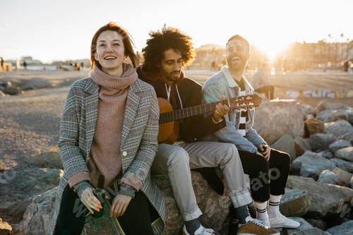 Preview: Three happy friends with guitar sitting outdoors at sunset