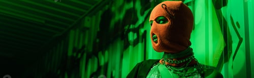 Preview: sexy anonymous woman in orange balaclava and silver necklaces looking away in green light near wall
