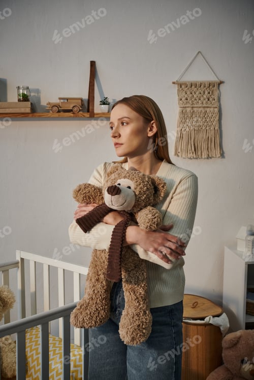 Preview: heavy-hearted grieving woman with soft toy standing near crib in bleak nursery room at home