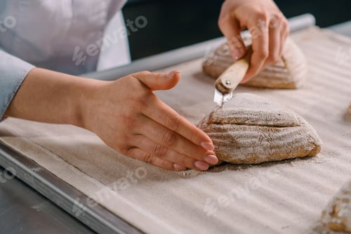 Preview: woman baker cuts patterns on raw bread rolls with a professional baker's knife before baking