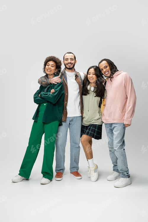 Preview: full length of happy multicultural students in trendy outfit smiling at camera on grey background