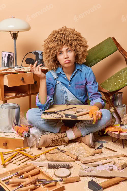 Preview: Hesitant curly haired female carpenter works in carpentry workshop works with wood repairs furniture