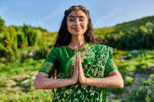 Preview: pleased young indian woman in green sari doing praying hands gesture with landscape on background