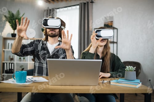 Preview: Excited young people with surprised expression, playing games at home, using VR glasses and laptops.