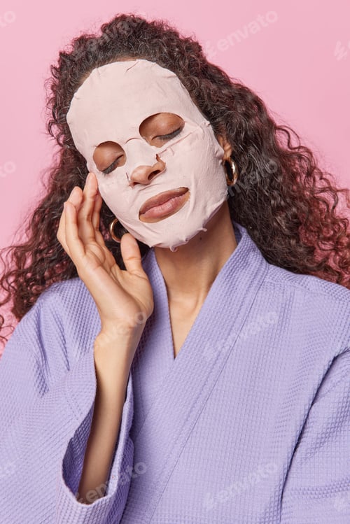 Preview: Vertical shot of calm charming young woman with long curly hair applies beauty sheet mask on fac for