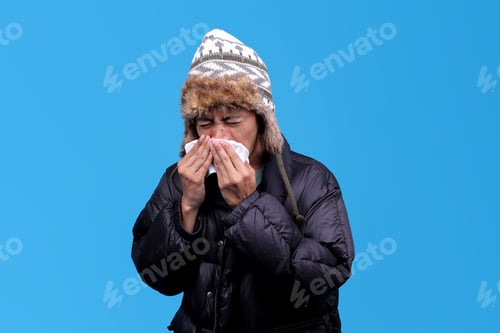 Preview: Man in winter clothes blowing his nose on a blue background, looking unwell