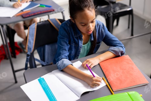 Preview: Happy biracial schoolgirl sitting at desk and taking notes in school classroom