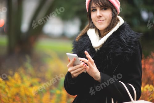 Preview: Young woman in a pink hat and winter coat using a smartphone on the street in autumn