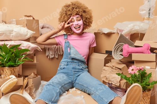 Preview: Positive carefree woman with curly hair makes peace gesture over eye applies beauty mask on face dre