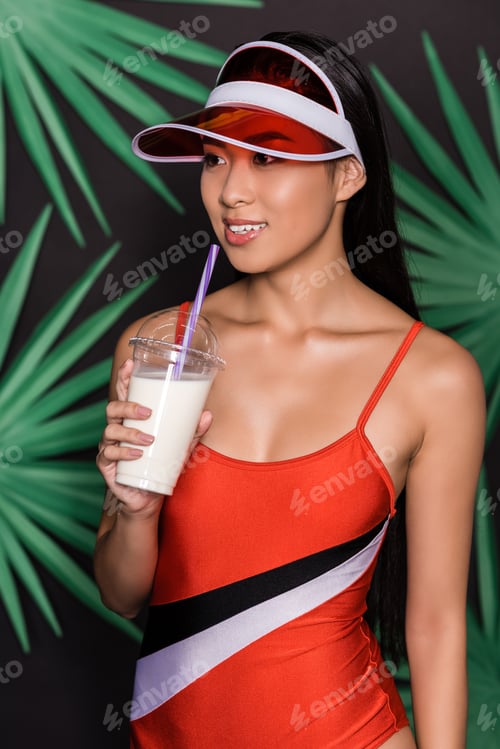 Preview: Half-length shot of beautiful asian woman in orange swimsuit and visor drinking milkshake and