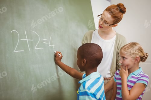 Preview: Side View Of Teacher Assisting Little Boy To Write On Blackboard In The Classroom